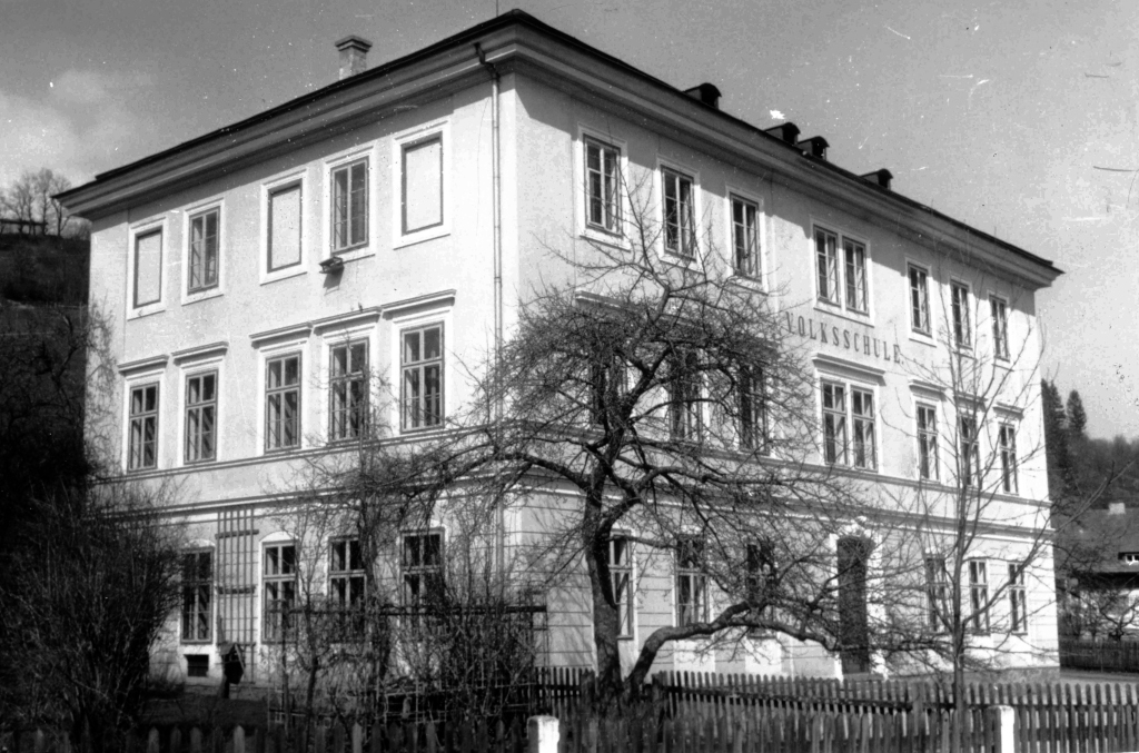 Ein dreistöckiges rechteckiges Gebäude mit hohen Flügelfenstern und der Aufschrift "Volksschule". Davor blattlose Obstbäume und ein Lattenzaun aus Holz.