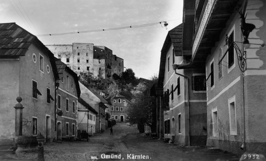 Bild der Kirchgasse in Gmünd nach Nordosten, vorne links ein Brunnen, Häuser säumen die Gasse, im Hintergrund auf dem Hügel die Burgruine