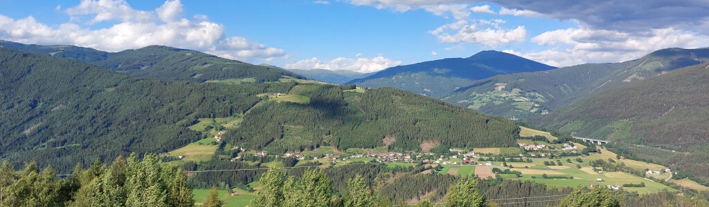 Blick von oben auf ein Plateau an einem bewaldetem Abhang mit einer Siedlung. Im Hintergrund bewaldete Bergkuppen. Im Vordergrund Laubbäume, die vom Wind nach rechts geneigt werden.